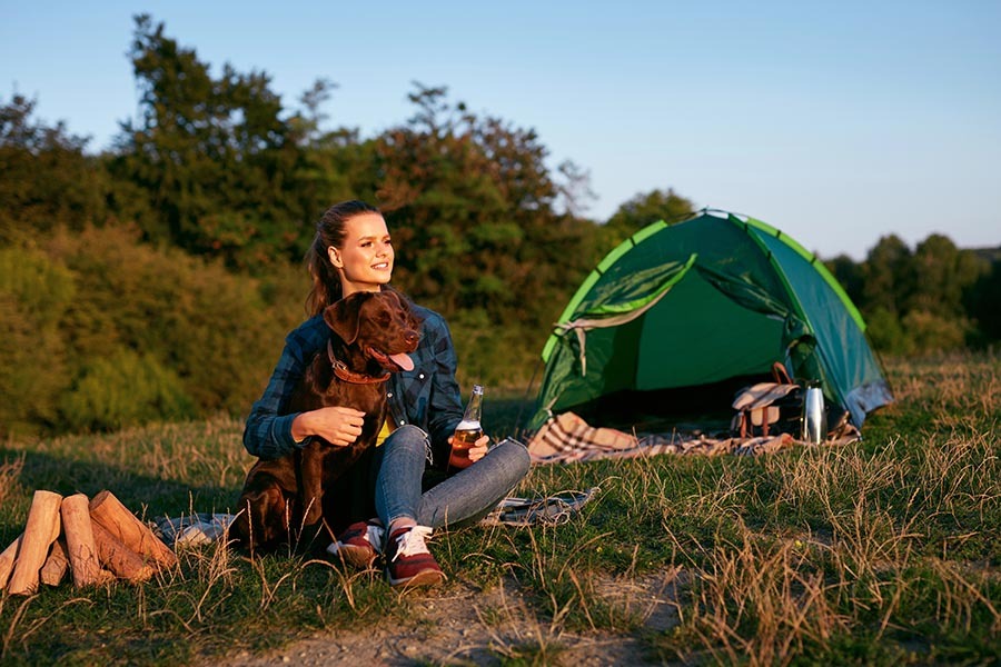 Happy Woman Traveling With Dog On Weekend In Nature. Beautiful Smiling Girl Sitting Near Camping Tent With Beer And Bonfire, Spending Time Together With Her Pet On Travel Vacation.